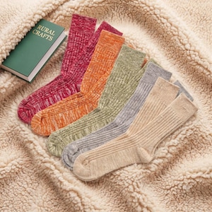 Puede incluir: Un conjunto de cinco calcetines acanalados en tonos magenta, naranja, verde oliva, gris y beige, dispuestos en abanico junto a un libro verde con el texto RURAL CRAFTS impreso en oro en la portada, todo sobre una tela de color crema texturizada.