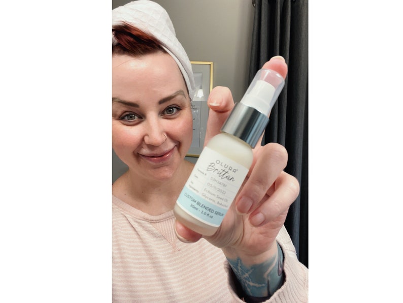 Puede incluir: Una botella blanca con una bomba de plata y una etiqueta azul que dice "OLURRA Brilliance" y "CUSTOM BLENDED SERUM 30ml | 1.01 fl oz".