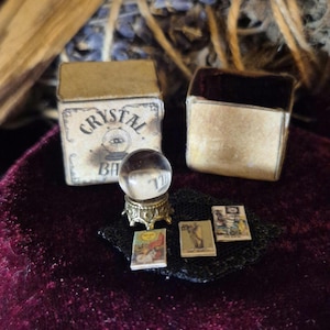 May include: A miniature crystal ball on a gold stand, with three tiny books and two small boxes. One box is labeled "Crystal Ball". The items are arranged on a dark red velvet surface, suggesting a mystical or fortune-telling theme.