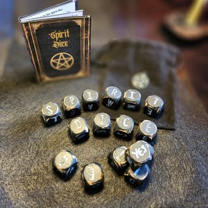 May include: Black spirit dice with white letters spelling out "Spirit Dice" on a dark brown surface. The dice are next to a small black book with gold trim and the words "Spirit Dice" on the cover.