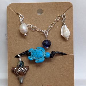 May include: Handmade jewelry set featuring a silver chain necklace with a pearl, a shell, and a dark blue bead charm. A black cord bracelet has a turquoise turtle charm and a shark tooth pendant. The card reads "Handmade with love."