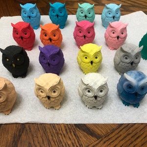 May include: A collection of 18 small owl figurines, each painted in a different color, including black, white, gray, blue, green, yellow, orange, pink, red, and purple. The owls are arranged in rows on a white surface.