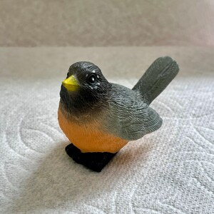 May include: A small, gray and orange bird figurine with a yellow beak. The bird is sitting on a white surface.