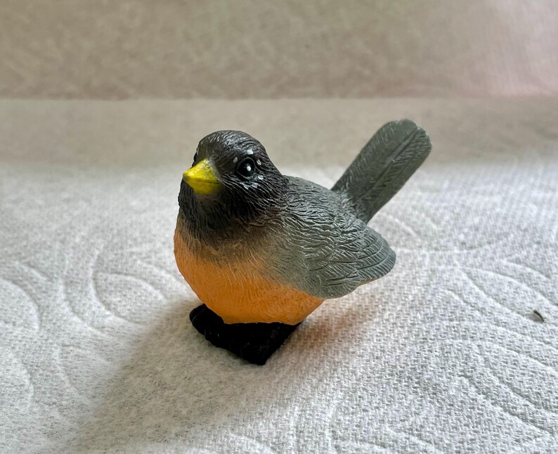 May include: A small, gray and orange bird figurine with a yellow beak. The bird is sitting on a white surface.