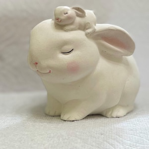 May include: White ceramic bunny figurine with a smaller white bunny figurine on its head. Both bunnies have closed eyes and pink noses.