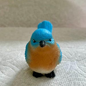 May include: A bluebird figurine with a brown chest and black eyes. The bird is looking directly at the camera.
