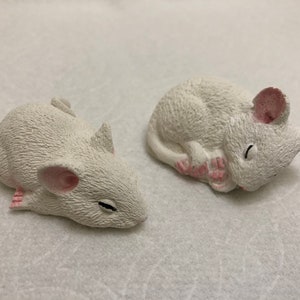 May include: Two white ceramic mice figurines, one lying on its side with its paws tucked under its chin, the other lying on its belly with its head resting on its paws.