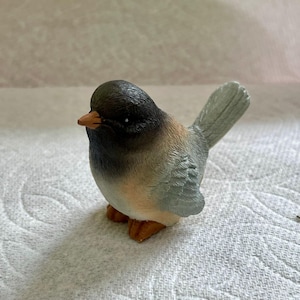 May include: A small, ceramic bird figurine with a black head, brown and gray body, and a light blue tail. The bird has a small, orange beak.