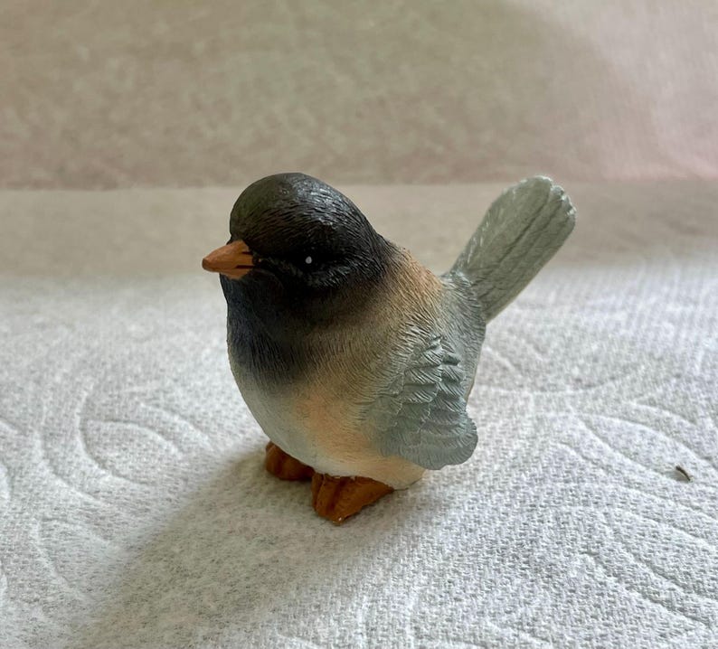 May include: A small, ceramic bird figurine with a black head, brown and gray body, and a light blue tail. The bird has a small, orange beak.