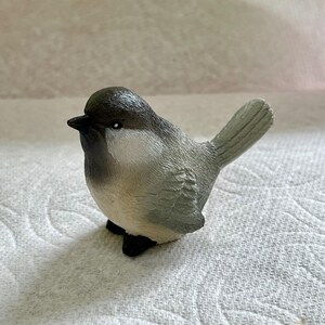 May include: A small gray and white bird figurine with a black eye. The bird is perched on a white surface.