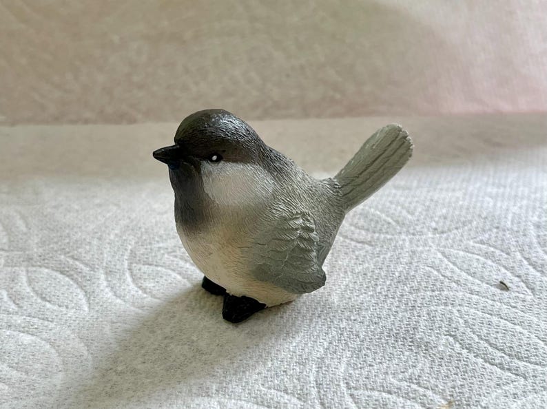 May include: A small gray and white bird figurine with a black eye. The bird is perched on a white surface.