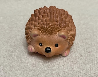 May include: A small brown hedgehog figurine with black eyes and a black nose. The figurine has a textured brown body and pink ears.