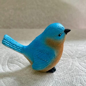 May include: A small blue bird figurine with a brown breast and black eyes. The bird is perched on a white surface.