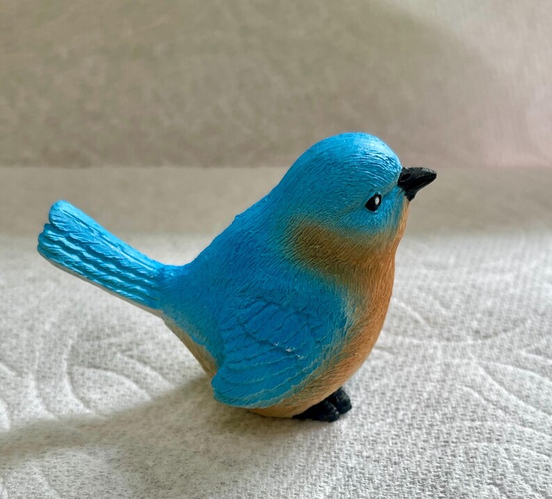 May include: A small blue bird figurine with a brown breast and black eyes. The bird is perched on a white surface.