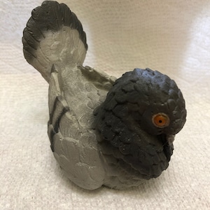 May include: A gray and black ceramic bird figurine with a detailed texture. The bird has a small orange eye and is sitting on a white surface.