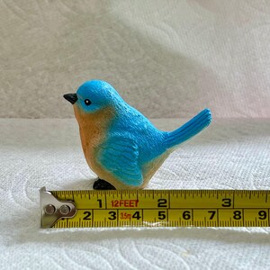 May include: A small bluebird figurine with a brown chest and black eyes. The figurine is sitting on a yellow measuring tape with the numbers 1, 2, 3, 4, 5, 6, 7, 8, 9, and 10 visible.