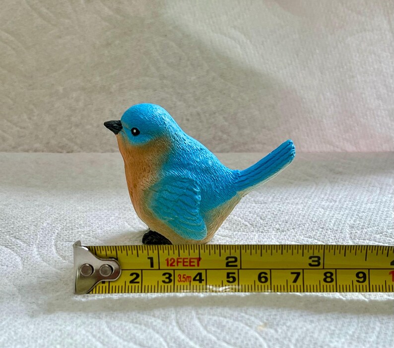 May include: A small bluebird figurine with a brown chest and black eyes. The figurine is sitting on a yellow measuring tape with the numbers 1, 2, 3, 4, 5, 6, 7, 8, 9, and 10 visible.