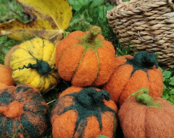 May include: A collection of handmade felt pumpkins in various shades of orange, green, and yellow, arranged on a bed of green grass. A woven basket sits in the background. The pumpkins vary in size and design, perfect for fall decor.