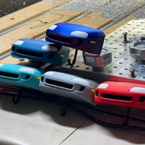 Puede incluir: Una colección de seis modelos de coches de juguete impresos en 3D en varios colores, incluyendo naranja, azul, verde azulado, gris y rojo. Los modelos son todos del mismo diseño, con una vista frontal de un coche con faros y una parrilla.