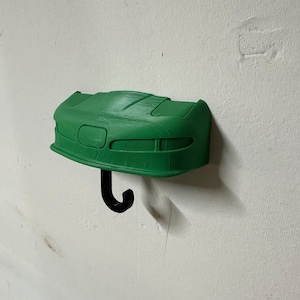 Puede incluir: Un gancho de pared de plástico verde en forma de la parte delantera de un coche. El gancho tiene la forma de la parte delantera de un coche deportivo con un gancho de metal negro.