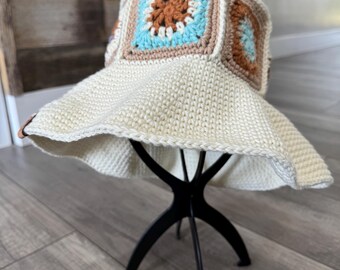 May include: A cream-colored crochet bucket hat with square, colorful accents. The squares feature a light blue center with a brown floral design, framed by tan and brown borders. The hat is displayed on a black stand.