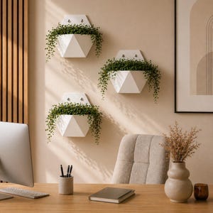 May include: Three white geometric wall planters, each filled with trailing green plants, are mounted on a beige wall. The planters have a modern, angular design. The scene includes a desk with a computer, a chair, and decorative items.