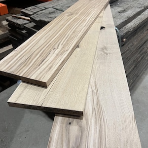 May include: Three long, rectangular wooden boards stacked in a staggered arrangement. The boards feature visible natural wood grain patterns in light brown and tan tones, with distinct knots and linear textures running along the length of each plank.