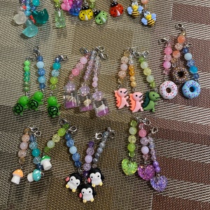 May include: A collection of colorful beaded keychains with various charms, including turtles, dinosaurs, donuts, ladybugs, bees, mushrooms, penguins, hearts, and glass bottles.