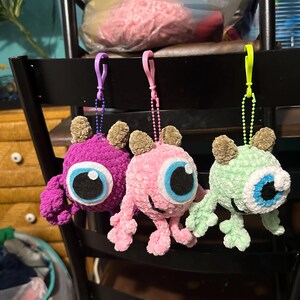May include: Three crocheted plush toys hanging from a chair. The toys are shaped like monsters with one large eye and are purple, pink, and green.