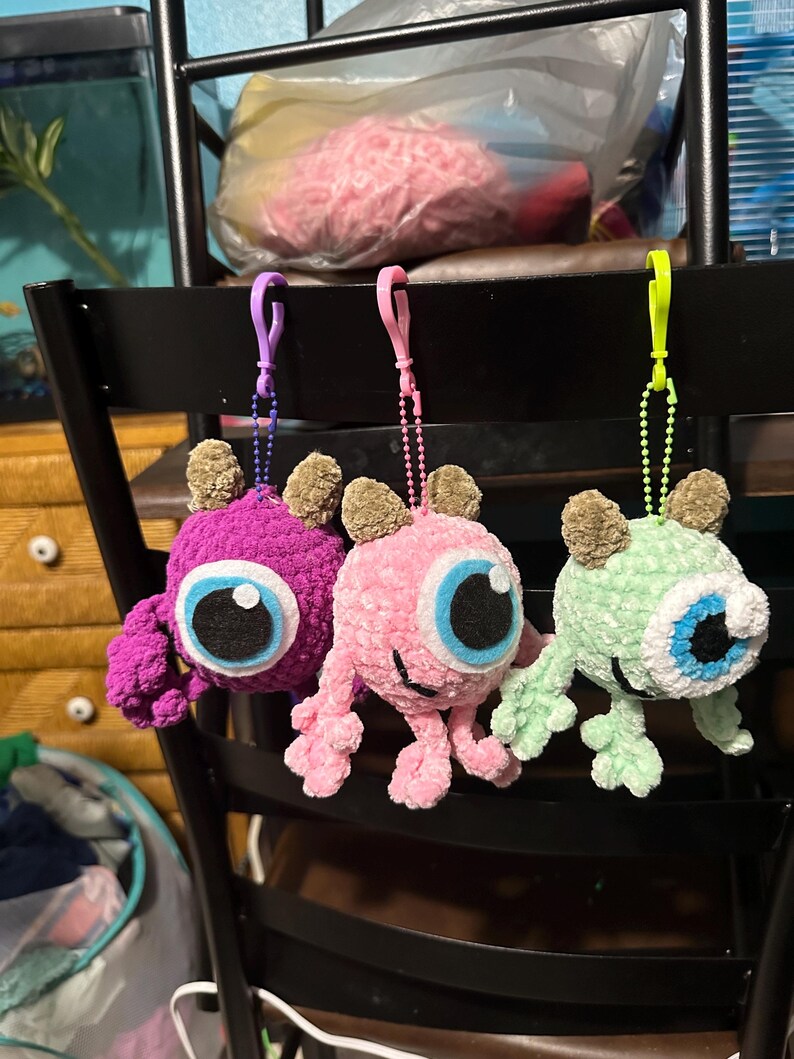 May include: Three crocheted plush toys hanging from a chair. The toys are shaped like monsters with one large eye and are purple, pink, and green.