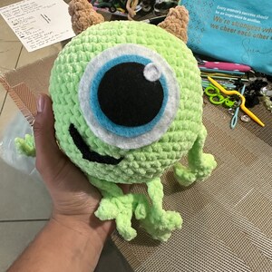 May include: A green crocheted plush toy with one large blue eye and a toothy grin. The toy is being held by a hand.