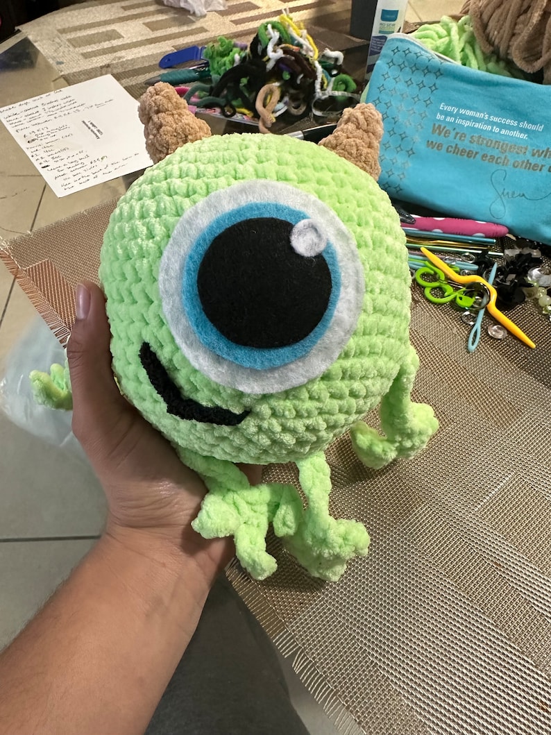 May include: A green crocheted plush toy with one large blue eye and a toothy grin. The toy is being held by a hand.