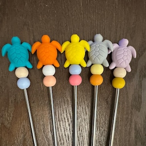 Puede incluir: Seis agitadores de bebidas con forma de tortuga de colores con pajitas de metal. Las tortugas son azules, naranjas, amarillas, moradas, grises y verdes. Las pajitas son plateadas.