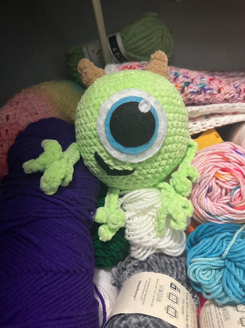 May include: A green crocheted monster toy with a large blue eye and a friendly smile. The toy is surrounded by balls of yarn in various colors, including purple, white, pink, and blue.
