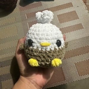 May include: A crocheted stuffed eagle toy with brown and white fur, yellow feet, and black eyes. The toy is being held in a person's hand.