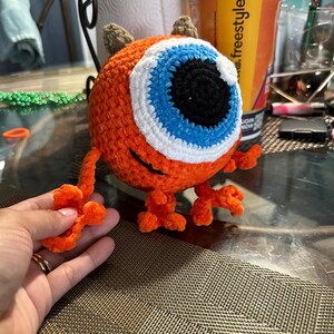 May include: A crocheted orange monster toy with a large blue eye, white eyelashes, and brown horns. The monster has orange legs and arms.