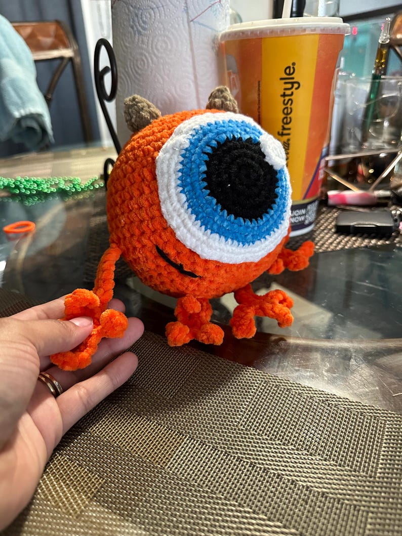 May include: A crocheted orange monster toy with a large blue eye, white eyelashes, and brown horns. The monster has orange legs and arms.