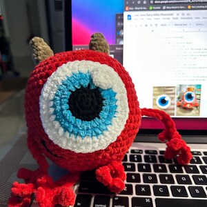 May include: A red crocheted monster with a large blue eye and white accents. The monster has two small horns and is sitting on a laptop keyboard.
