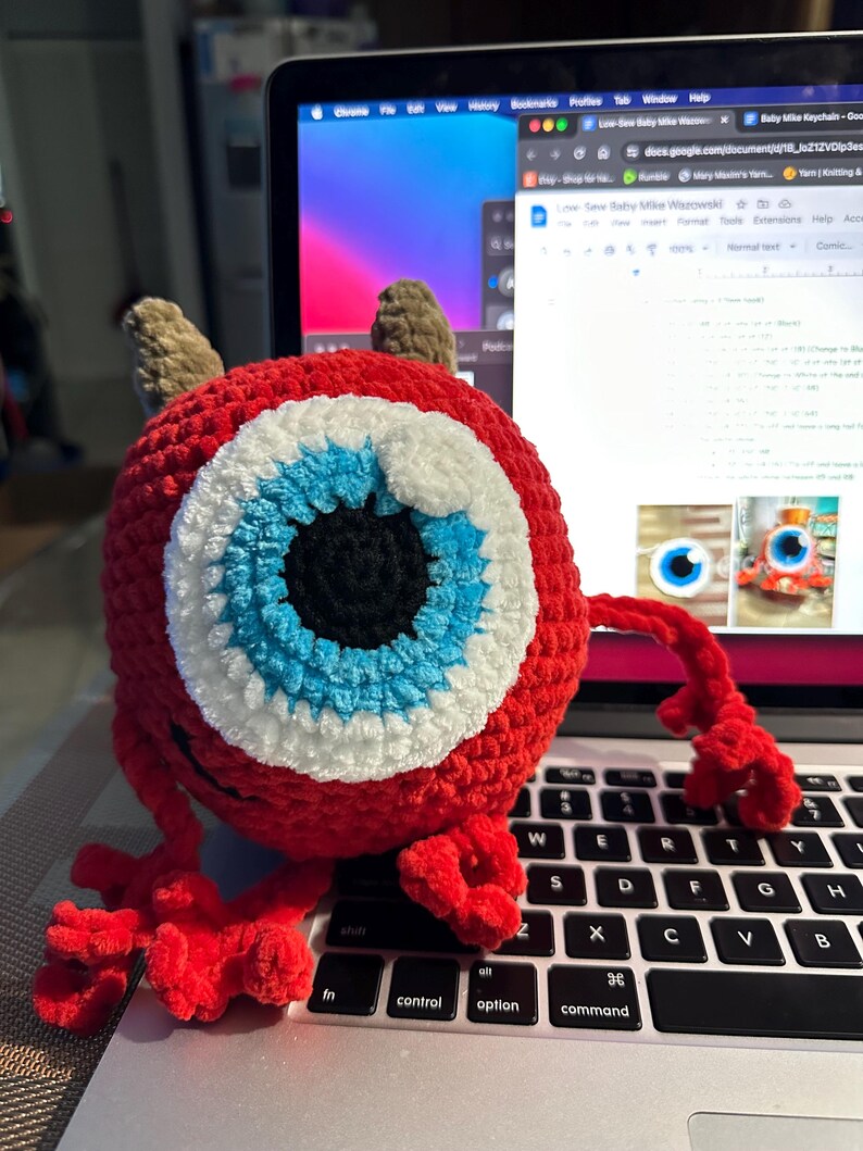 May include: A red crocheted monster with a large blue eye and white accents. The monster has two small horns and is sitting on a laptop keyboard.