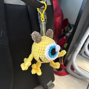 May include: A yellow crocheted plush toy with one large blue eye and brown ears hanging from a yellow plastic clip on a black backpack.