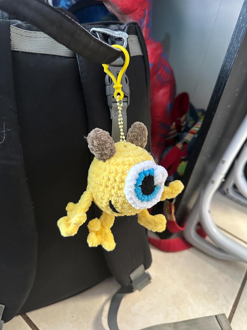 May include: A yellow crocheted plush toy with one large blue eye and brown ears hanging from a yellow plastic clip on a black backpack.