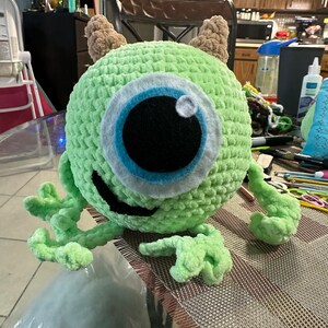 May include: A green crocheted plush toy with one large blue eye and a toothy grin. The toy has two brown yarn horns on top of its head.