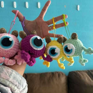 May include: Four crocheted plush toys with large eyes and different colors: pink, purple, yellow, and green. Each toy is attached to a keychain.