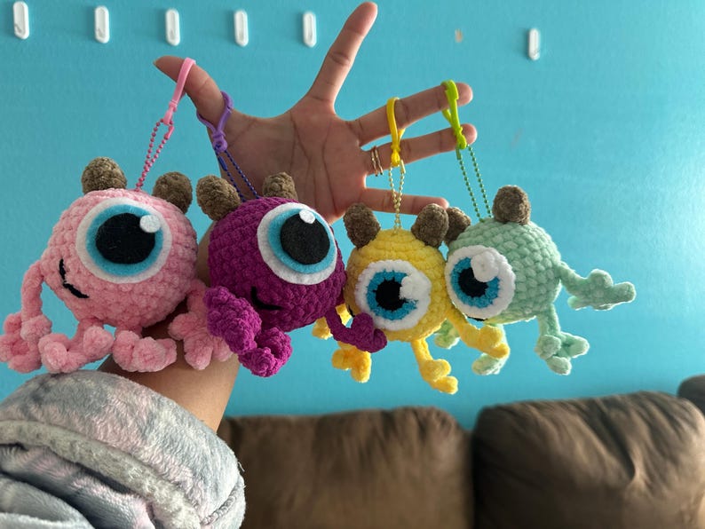 May include: Four crocheted plush toys with large eyes and different colors: pink, purple, yellow, and green. Each toy is attached to a keychain.