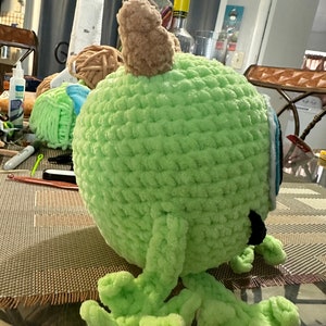 May include: A green crocheted plush toy with brown horns and blue eyes. The toy has four legs and is sitting on a brown surface.