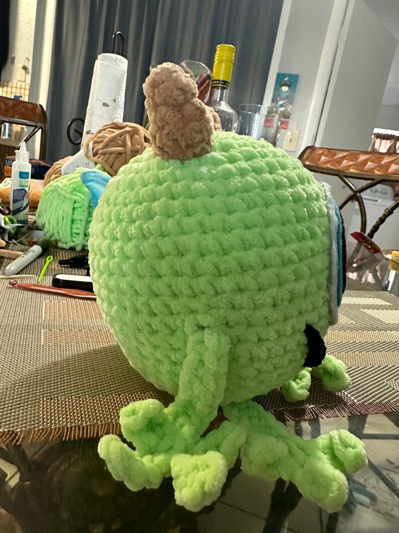 May include: A green crocheted plush toy with brown horns and blue eyes. The toy has four legs and is sitting on a brown surface.