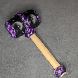 May include: A black and purple mallet with a wooden handle. The mallet head is made of black leather and is wrapped with purple and black rope. The handle is made of light-colored wood, and the base is also wrapped with the same rope.