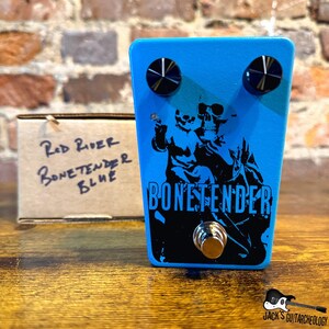 May include: A blue Bonetender guitar effects pedal with black knobs and a silver footswitch. The pedal features a black graphic of two skulls and the text "BONETENDER". A cardboard box with handwritten text is to the left.