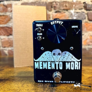 Könnte beinhalten: Schwarzes Gitarreneffektpedal mit dem Text "MEMENTO MORI" und einer Totenkopf-Grafik mit Flügeln. Verfügt über Knöpfe für Höhen, Bässe, Drive und Output. Hergestellt von Red River Amplification.