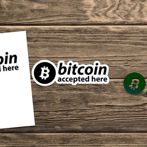 May include: Three stickers with the text "bitcoin accepted here" on a wooden background. The stickers feature the Bitcoin logo and text in black and green. One sticker is on a white background.